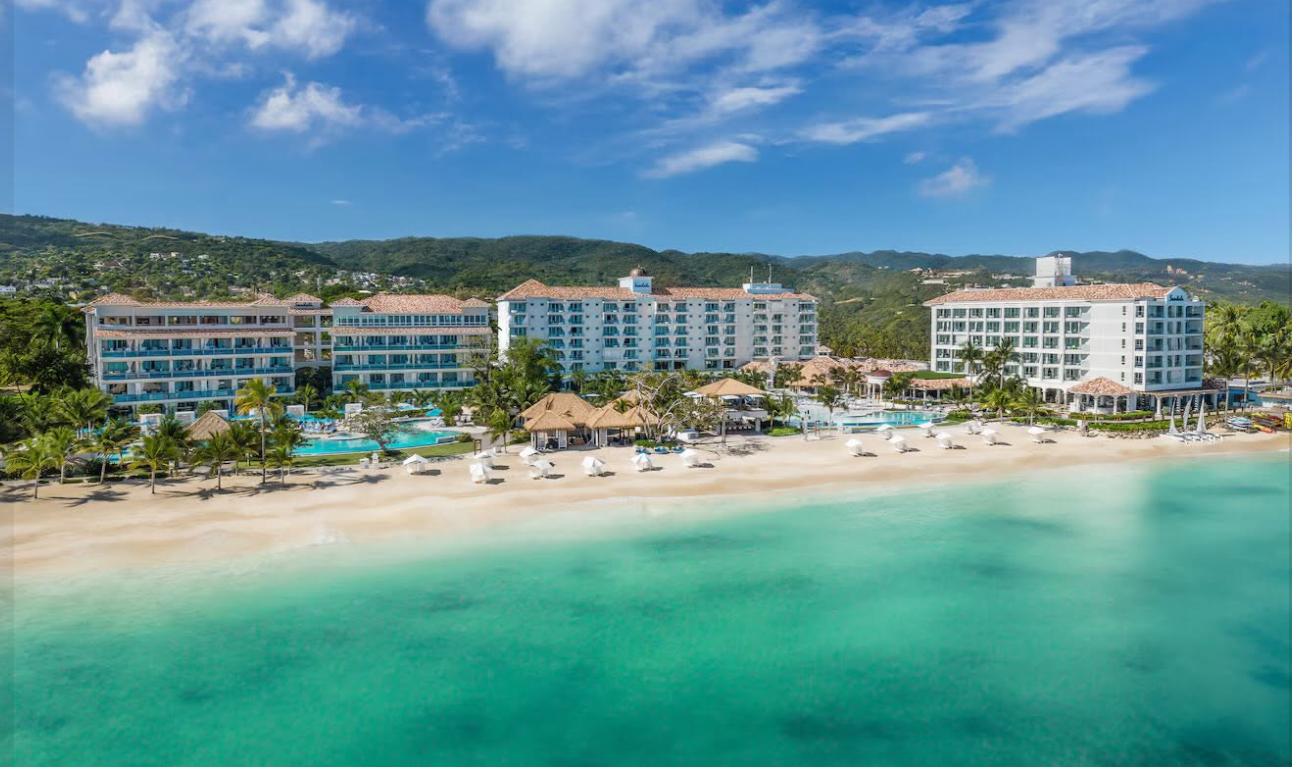 Beachfront view of Sandals Dunn's River in Jamaica: resort buildings and pools behind a wide sand beach and turquoise Caribbean water