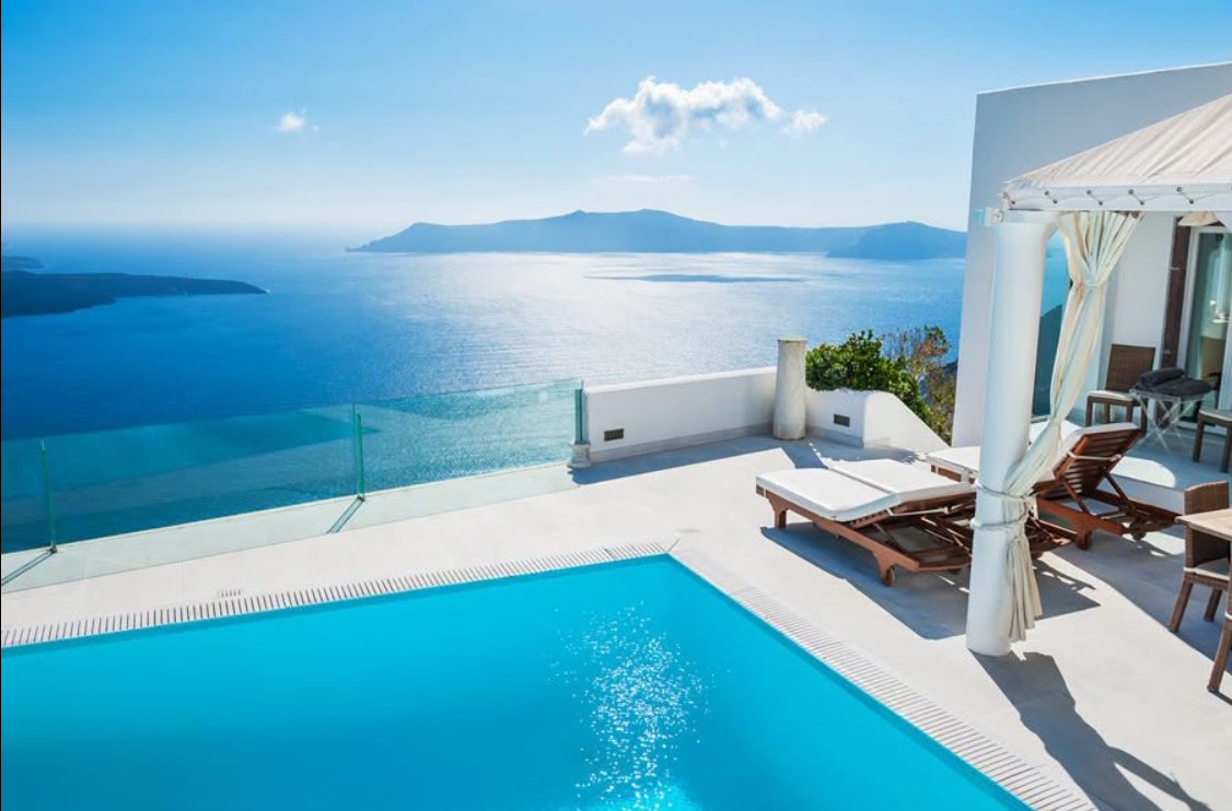 Whitewashed Santorini terrace with a bright blue infinity pool and loungers overlooking the Aegean caldera